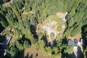 An aerial view of a forested area with a small cluster of buildings, including a house with a garage and a shed.