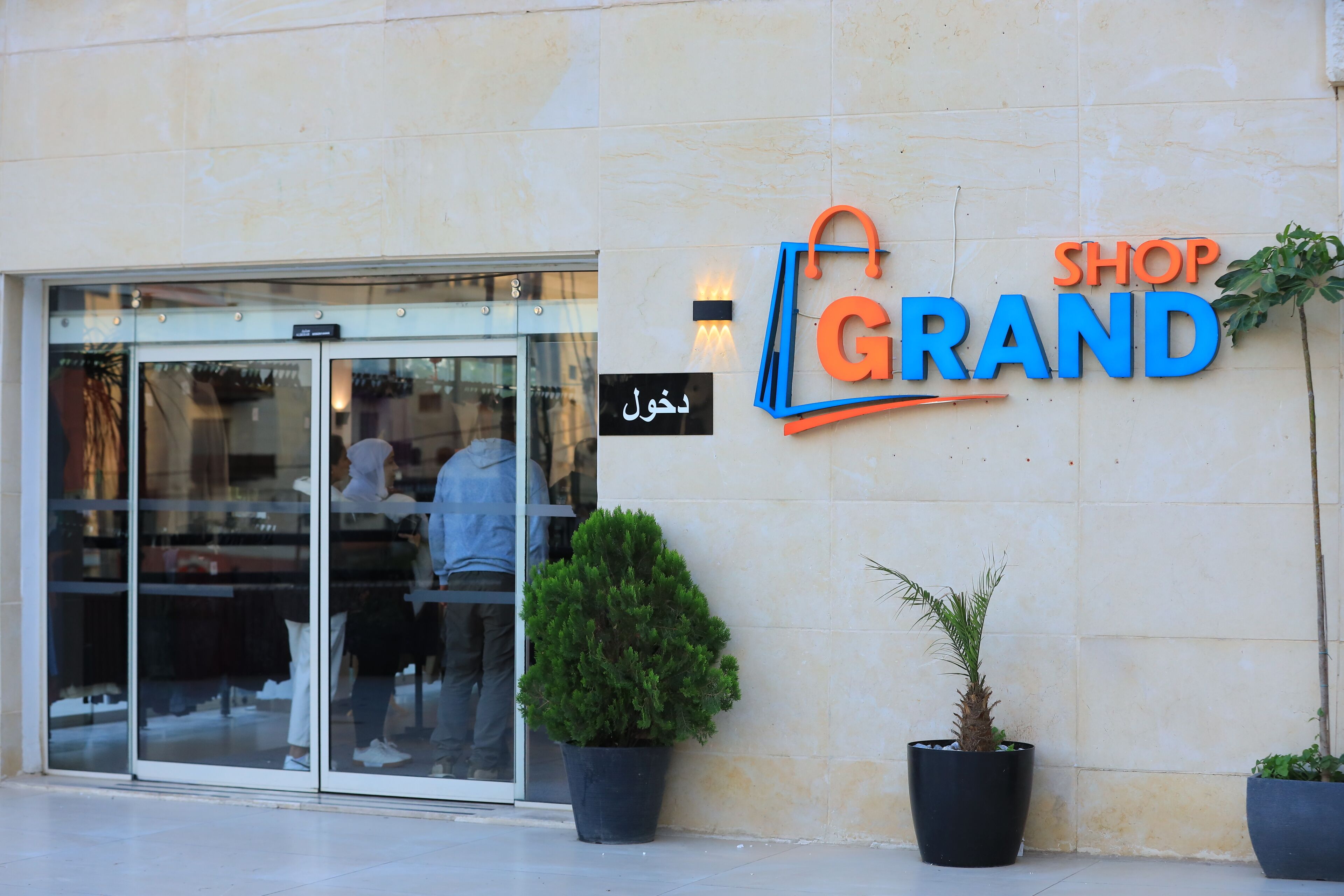 Une devanture de magasin avec une porte d’entrée en verre, une enseigne indiquant « MAGASIN », et deux plantes en pot de chaque côté de l’entrée.