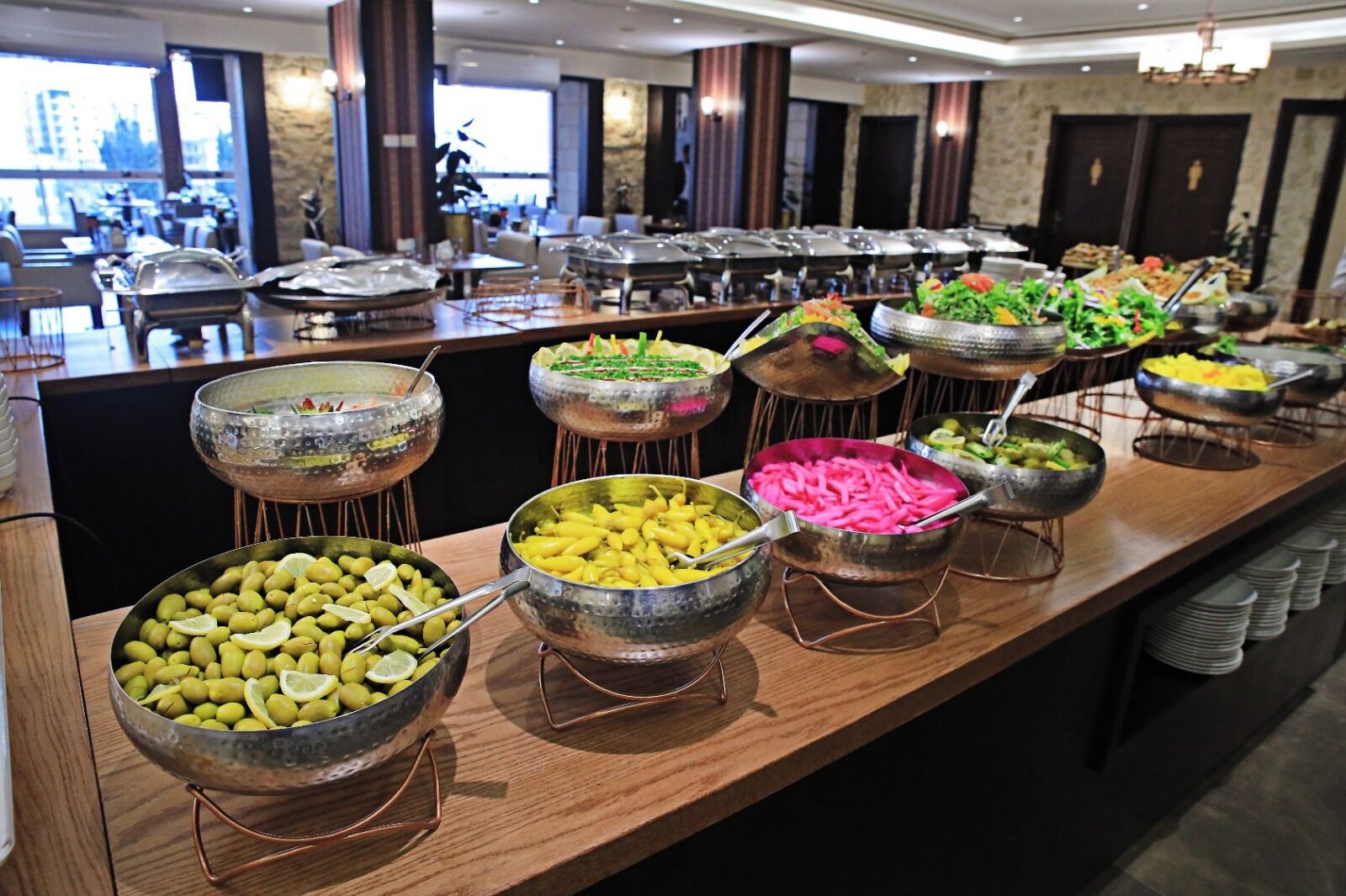 A buffet with various food items in metal bowls, a wooden counter, and a view of the city through large windows.