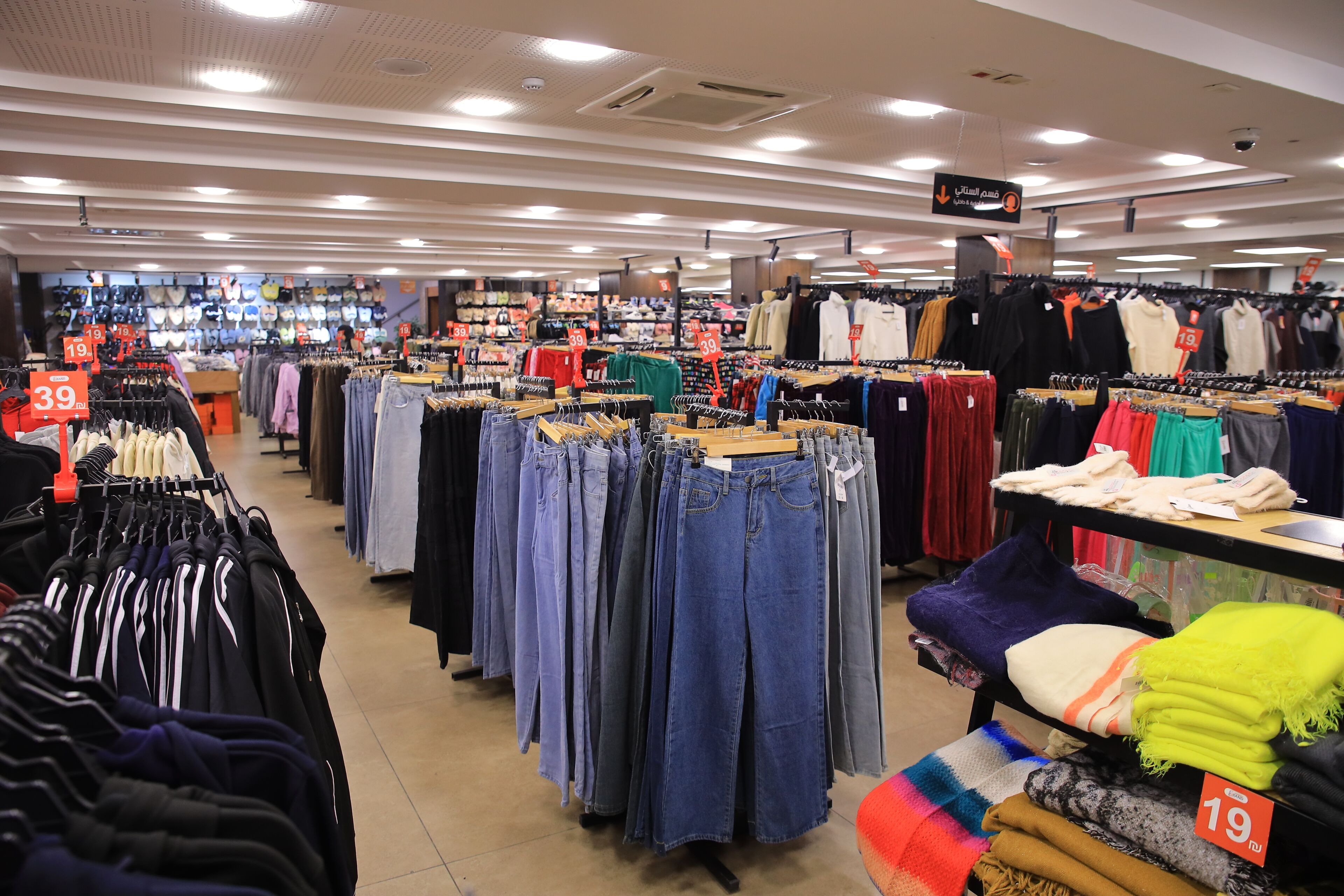 Tienda de ropa con estantes llenos de vaqueros y camisetas, etiquetas de precio y una gran variedad de prendas.