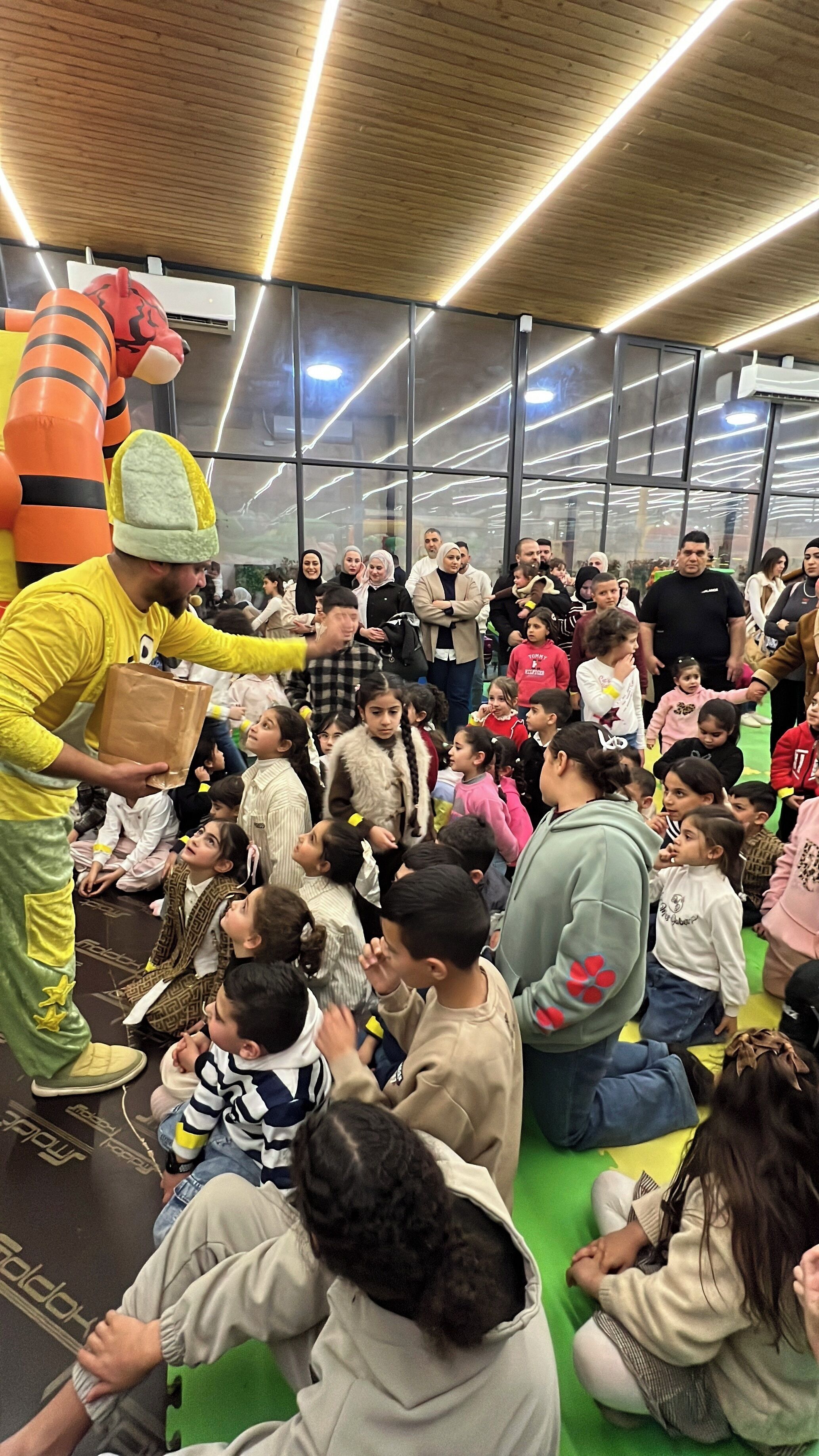 A group of people seated on a green floor, with a person in a costume holding a brown bag.