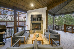 A covered outdoor seating area with a stone fireplace, wooden deck, and a view of trees.