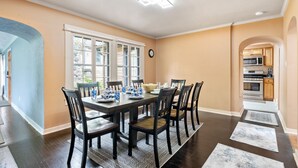 Un comedor con una gran mesa dispuesta para seis personas, rodeada de sillas. Hay un ventanal con múltiples vidrios, un reloj en la pared y una cocina visible a través de un arco.