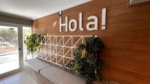 A reception area with a wooden wall, a 'Hola!' sign, a white grid partition, and potted plants.