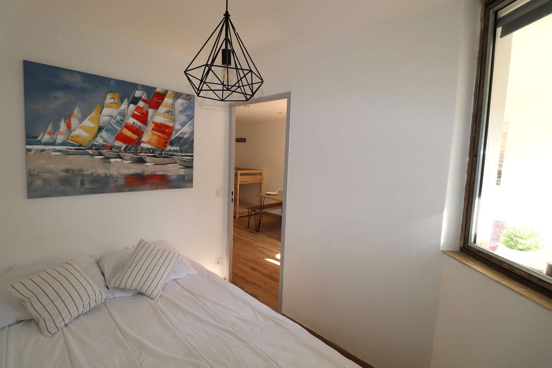 Une chambre à coucher avec un lit, un tableau représentant des voiliers, un luminaire aux formes géométriques et une fenêtre.