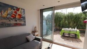 Een moderne woonkamer met een bank, een schilderij van zeilboten en een glazen schuifdeur die leidt naar een buitenruimte met een tafel en stoelen.