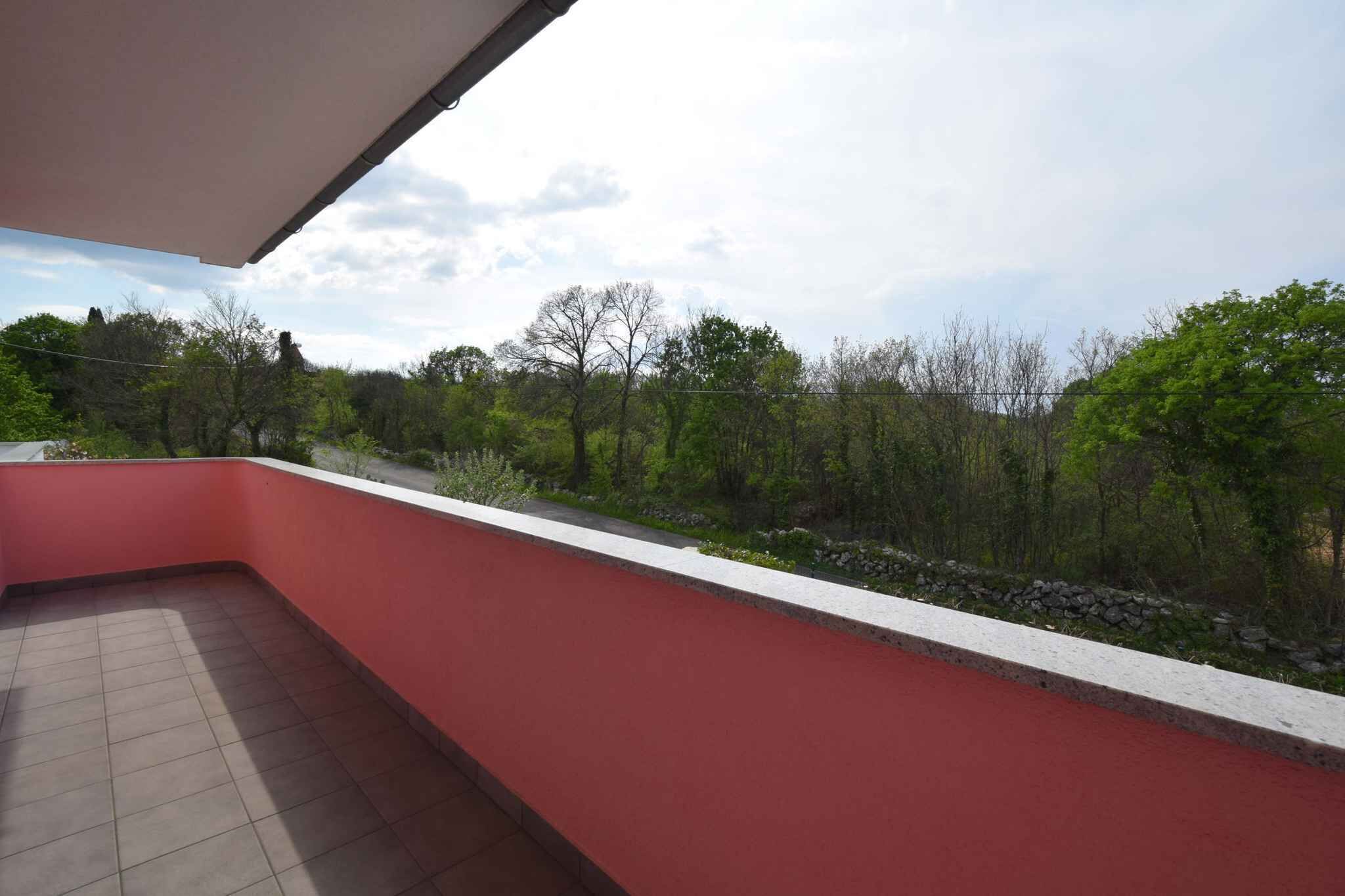 A red balcony with a tiled floor and a view of a landscaped area with trees and a stone wall.