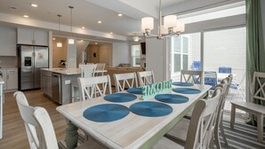 A modern kitchen with stainless steel appliances, a dining area with a wooden table and blue placemats, and a view of the outdoors through large windows. - Recently Built  Rehoboth Beach Townhouse Pool Parking Sleeps 16 (Rehoboth Beach)