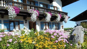 Un chalet traditionnel doté de balcons ornés de jardinières suspendues, entouré d’un jardin luxuriant où poussent différentes espèces de plantes à fleurs.