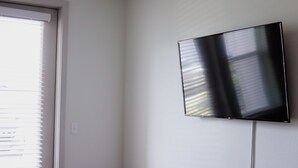 Une pièce dotée d’une grande fenêtre avec stores, d’un téléviseur à écran plat fixé au mur et d’une porte blanche.