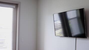 Huoneessa on suuri ikkuna, jossa on sälekaihtimet, seinälle asennettu taulutelevisio ja valkoinen ovi.