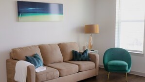Un canapé beige agrémenté d’un plaid blanc et de coussins turquoise, un fauteuil turquoise aux pieds dorés, un lampadaire et un tableau représentant une scène de plage.