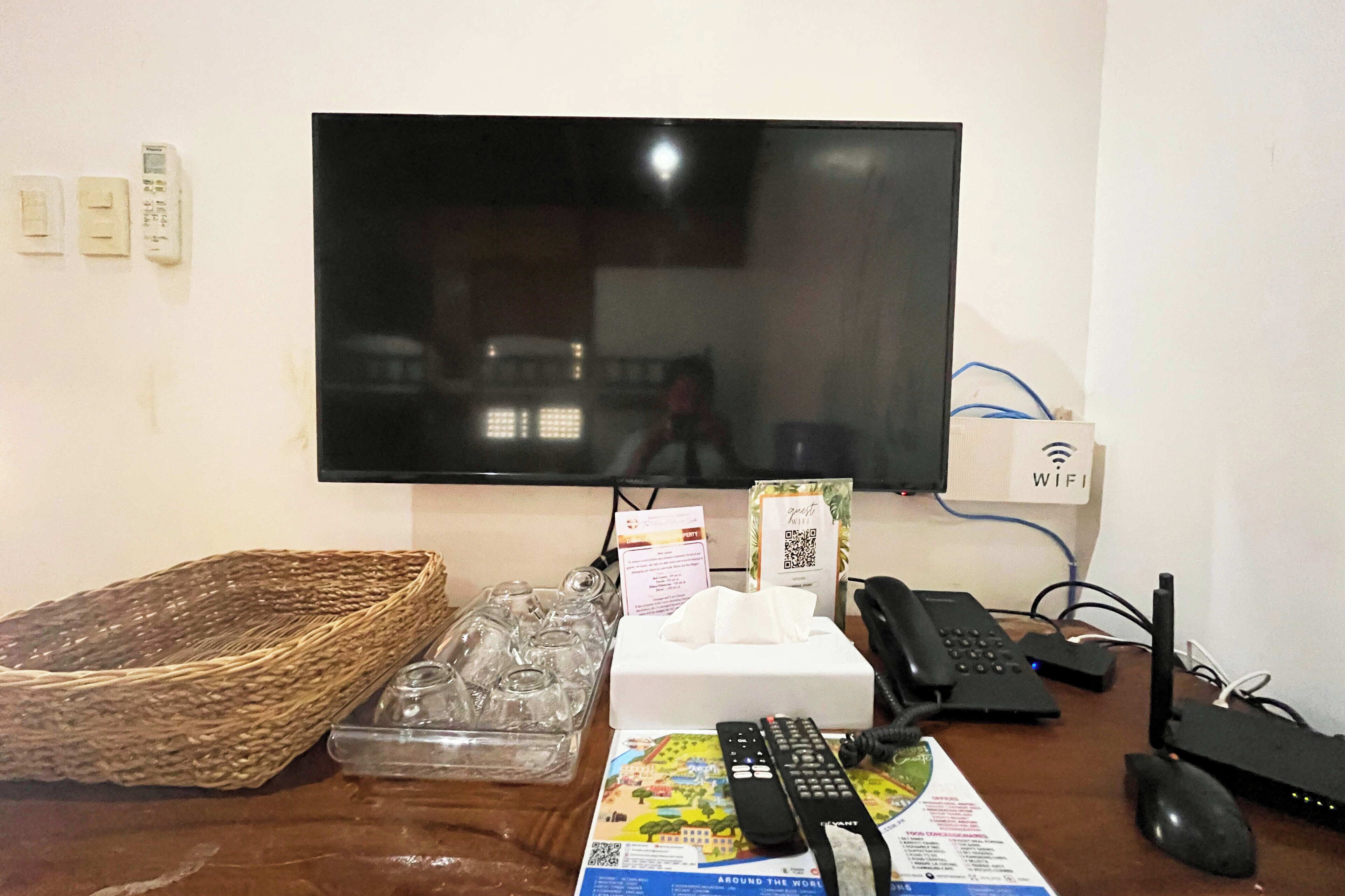 A desk with a flat-screen TV, a phone, a router, and a basket of glasses.
