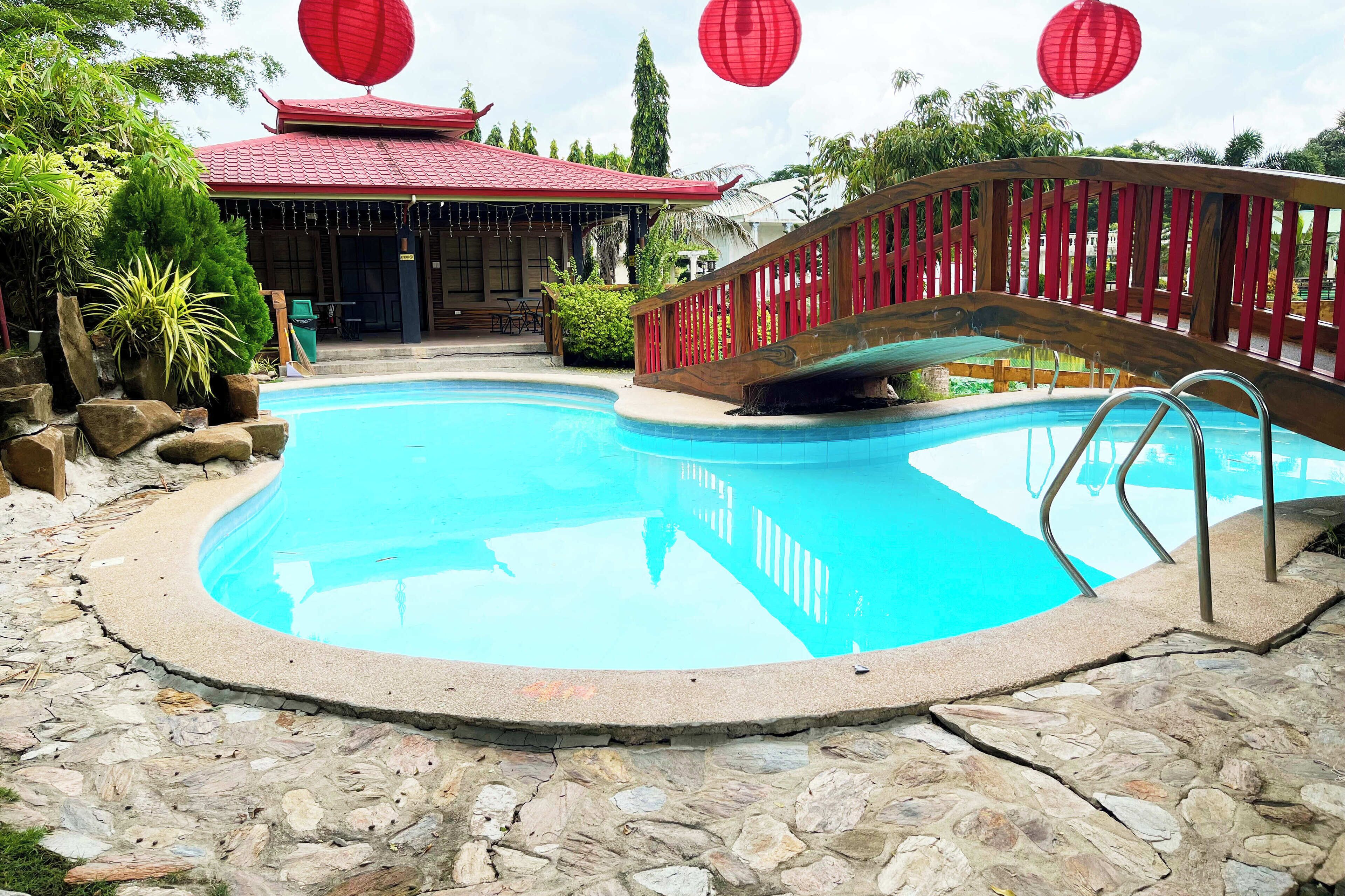 A pool area with a stone pathway, a wooden bridge, and a building with a red roof.