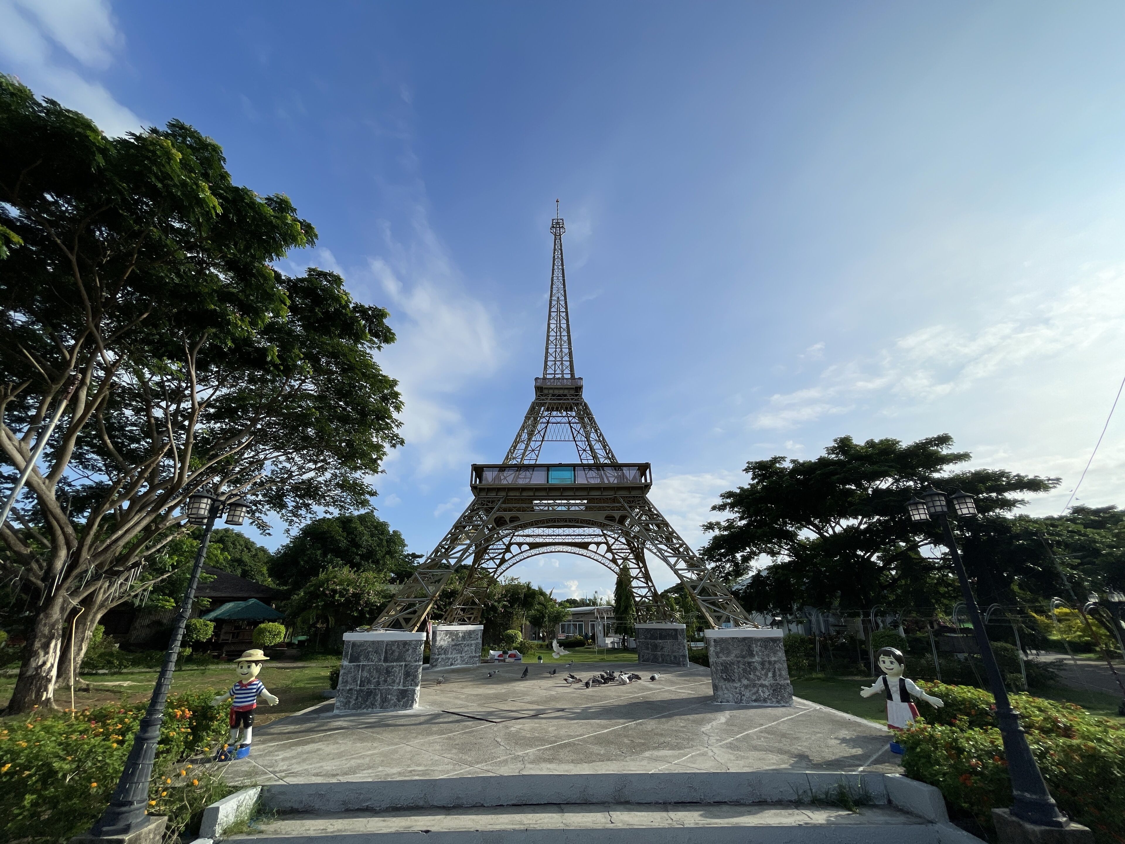 A model of the Eiffel Tower in a park with stone pillars, trees, and a statue of a man.