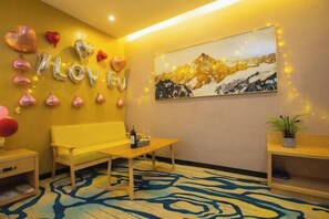 A room with a yellow sofa, a wooden table with a bottle, balloons spelling 'LOVE YOU,' and a mountain landscape painting.