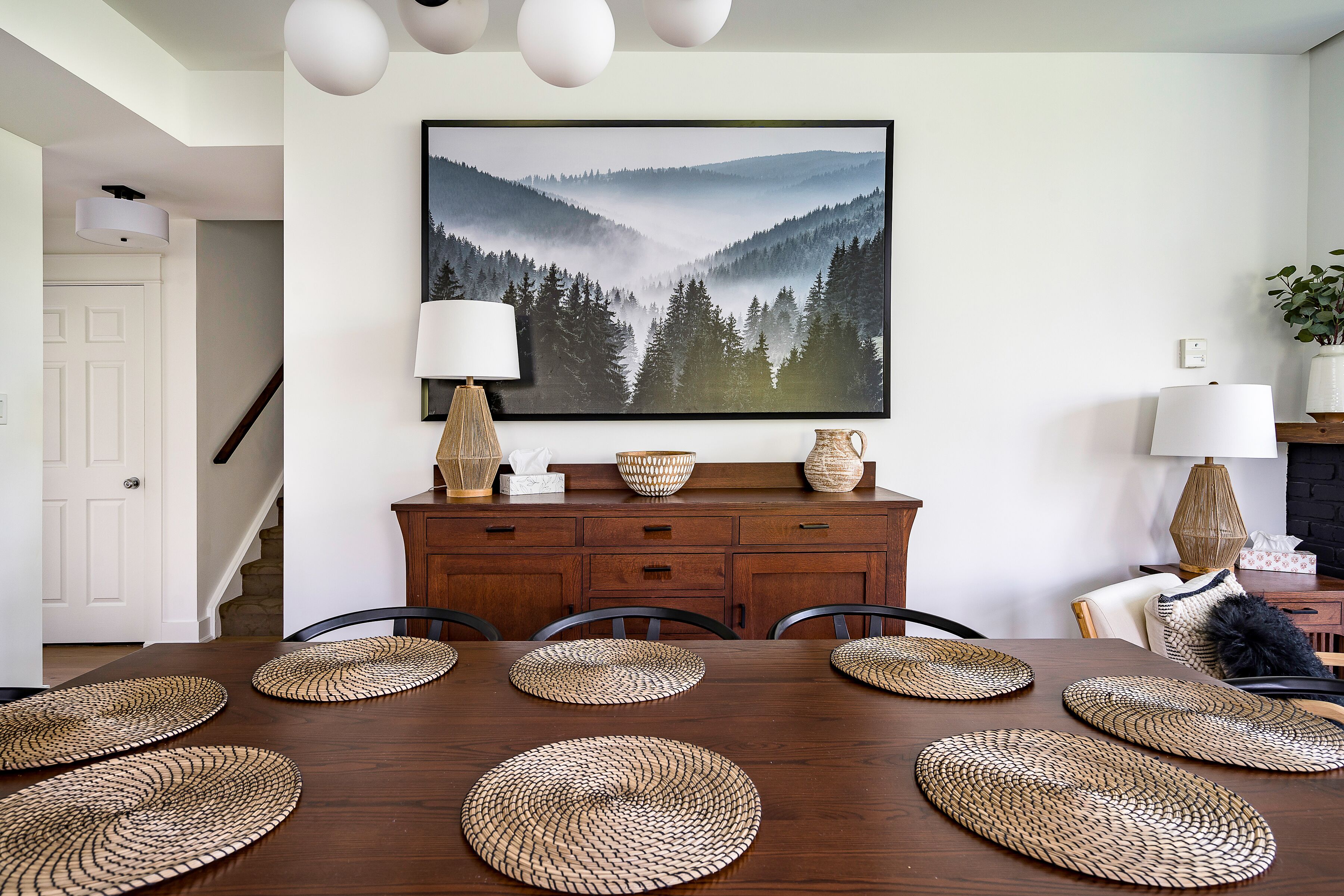 Comedor con mesa de madera, individuales tejidos y un cuadro grande de un paisaje forestal.