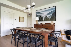 A dining area with a wooden table, black chairs, woven placemats, a large framed picture of a forest, and a lamp on the table.