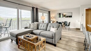 A modern living room with a grey sectional sofa, a glass-top coffee table, and a dining area with a table and chairs. - ★Gulf Coast 107★ Heated Pool + Beach Gear, Steps to Beach! Clean&Spacious Condo (Indian Shores)