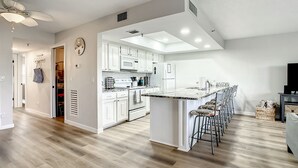 A modern kitchen with white cabinets, a central island with bar stools, and a ceiling fan. - ★Gulf Coast 107★ Heated Pool + Beach Gear, Steps to Beach! Clean&Spacious Condo (Indian Shores)