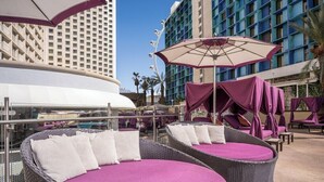 Un espace de détente aménagé sur le toit, avec des fauteuils en osier et des coussins violets, offrant une vue sur une piscine et des immeubles de grande hauteur.