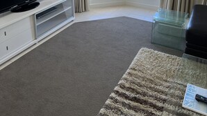 A living room with a flat-screen TV on a white entertainment unit, a glass coffee table, a beige rug, and two windows with curtains. - Enjoy a Break Away in This Lovely Home With Great Views (Middleton)
