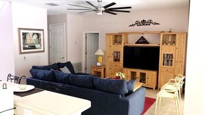 Ein Wohnzimmer mit einem dunkelblauen Sofa, einem Flachbildfernseher, Holzschränken und einem Deckenventilator.