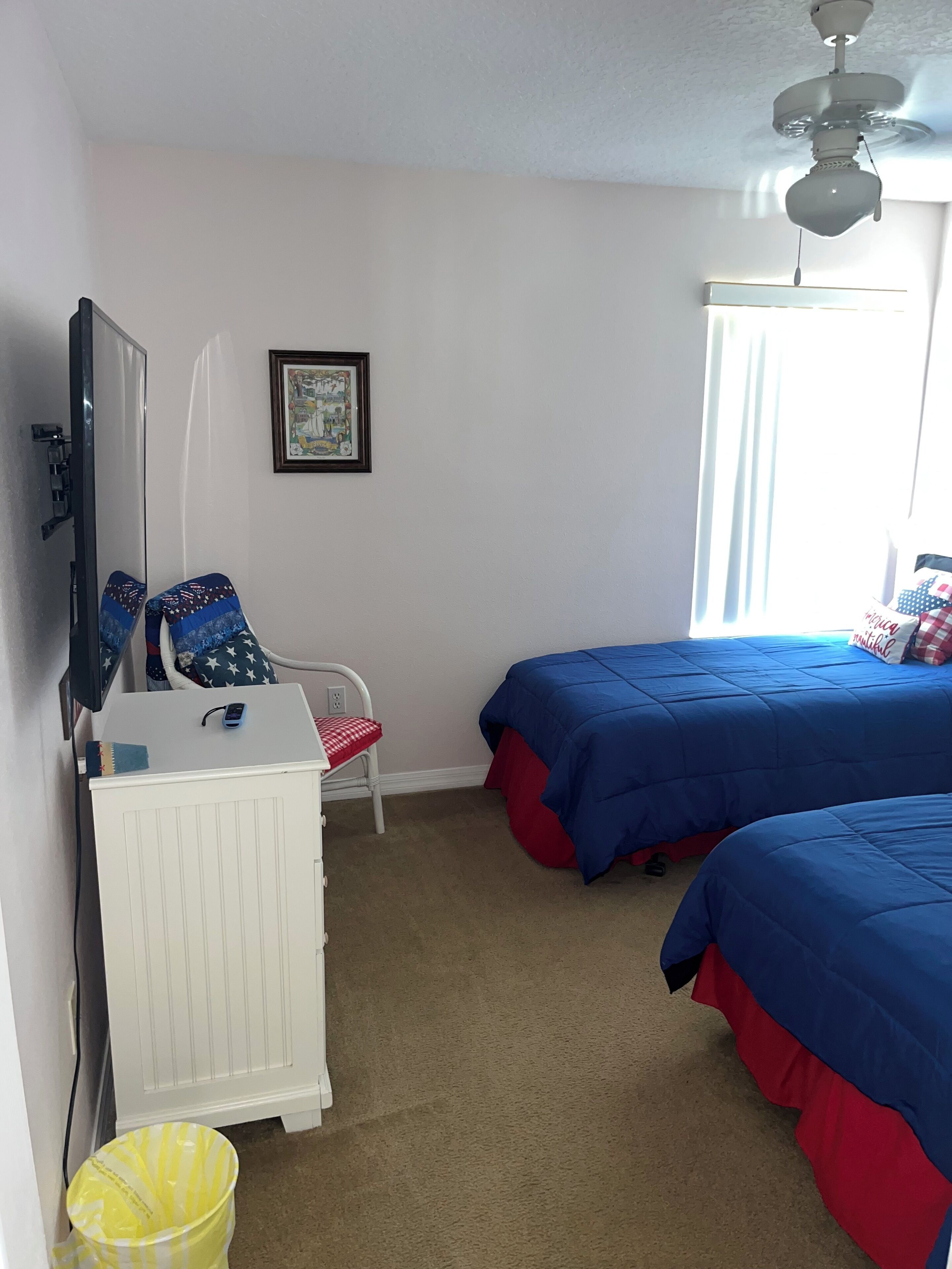 Een slaapkamer met twee bedden, een witte dressoir, een stoel, een plafondventilator en een raam met gordijnen.