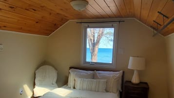 Une chambre avec un plafond en pente, un plancher en bois, un lit avec une literie blanche, une table de chevet avec une lampe et une fenêtre donnant sur la mer.