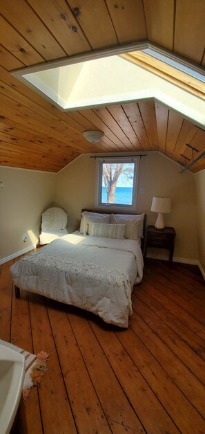A bedroom with a sloped ceiling, wooden flooring, a bed with white bedding, a nightstand with a lamp, and a window with a view of the sea.