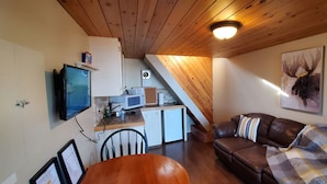 Un espace de vie compact comprenant un coin cuisine, un canapé en cuir marron, une table à manger avec des chaises et un téléviseur fixé au mur.