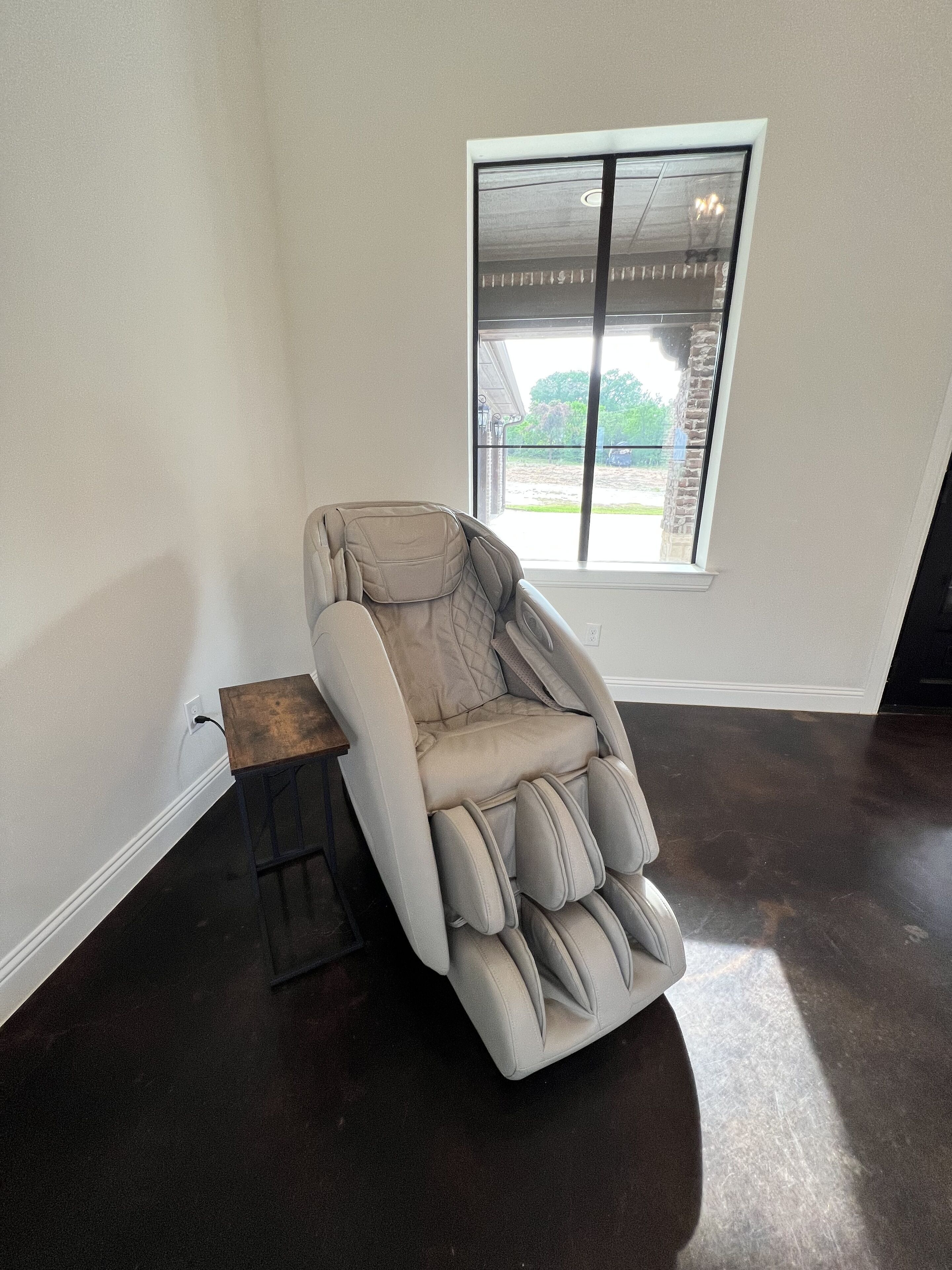 Una silla de masaje en una habitación con un ventanal grande, una mesita de madera y un suelo oscuro.