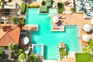 Ein großer Pool im Freien mit mehreren Liegebereichen und Sonnenschirmen, umgeben von gepflegter Bepflanzung und einer Dachkonstruktion mit Fliesen.