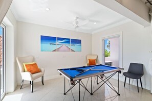 Ein Zimmer mit Billardtisch, zwei Sesseln, Deckenventilator und großem Fenster mit Meerblick.