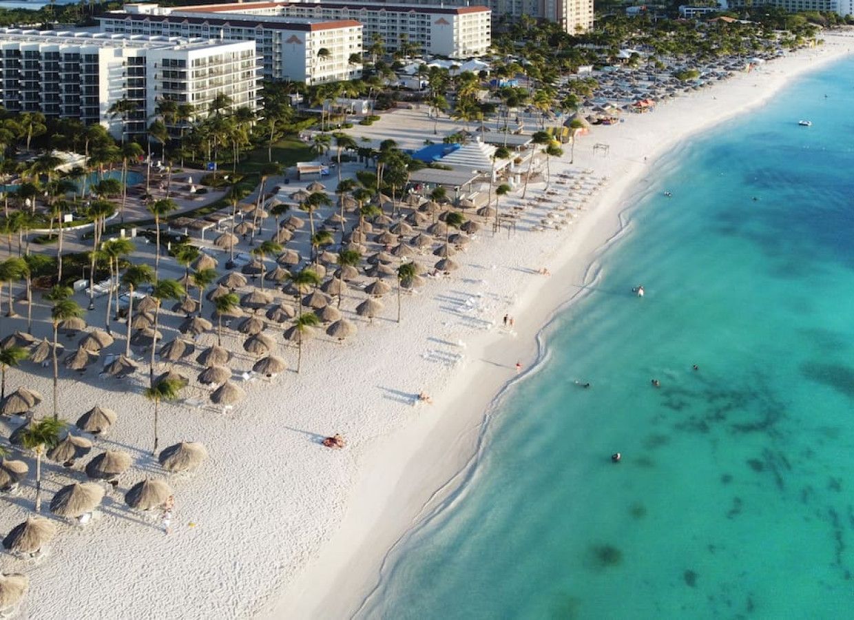 A beach resort with multiple hotel buildings, a sandy beach lined with palm trees and thatched umbrellas, and clear turquoise waters.