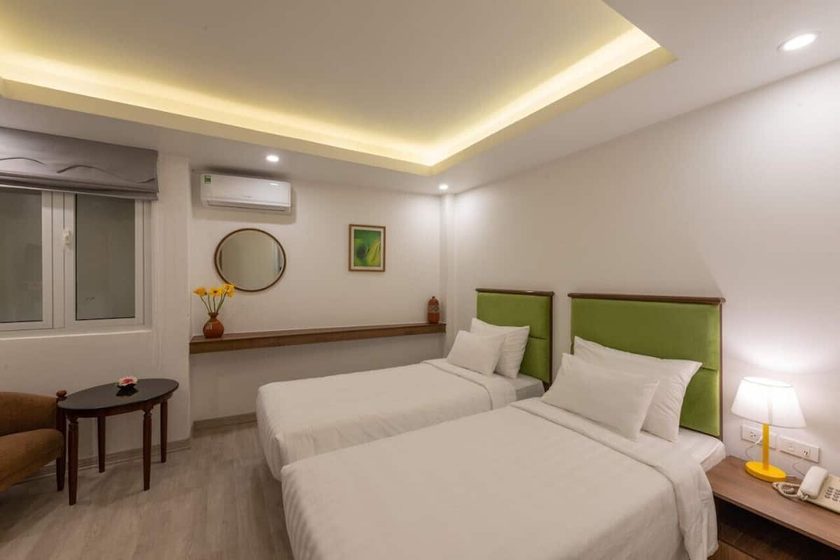 A hotel room with two beds, a small table, a chair, a bedside lamp, and a window with a view.