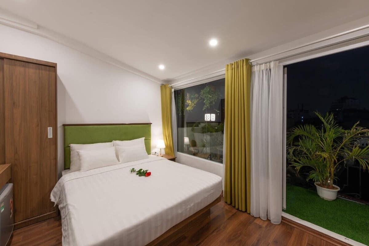 A hotel room with a bed, a green headboard, a nightstand, a window with curtains, and a potted plant.