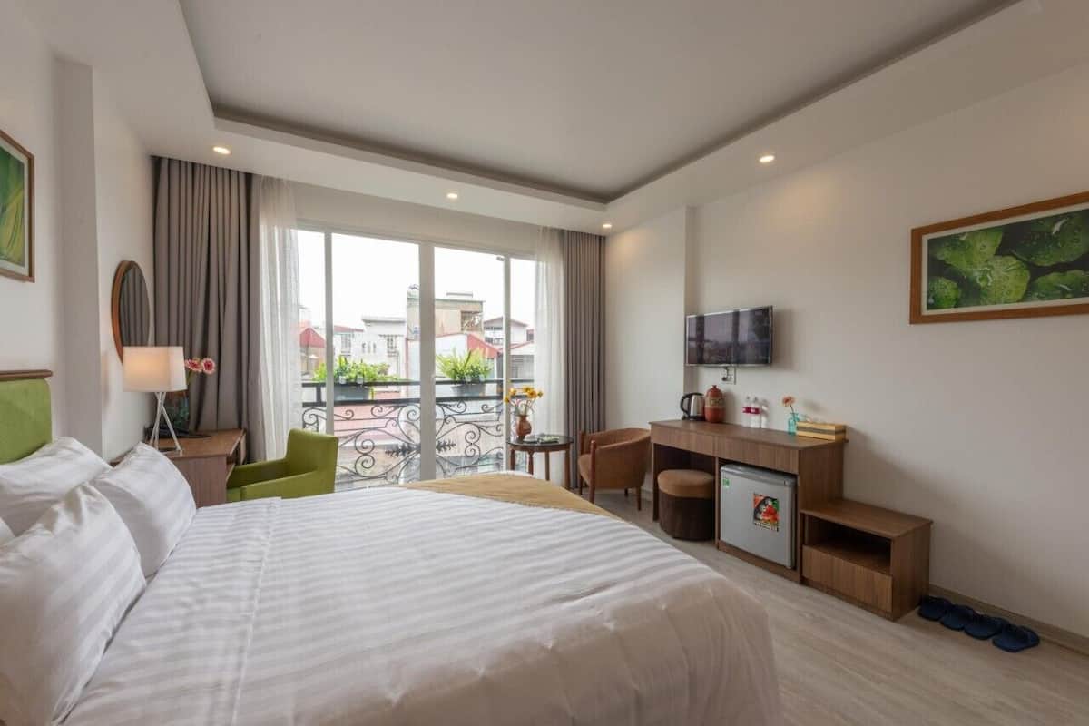 A hotel room with a large bed, a desk, a chair, a television, and a balcony with a view of buildings.