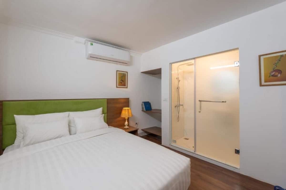 A hotel room with a bed, a green headboard, a bedside table with a lamp, a wardrobe, and an air conditioning unit.
