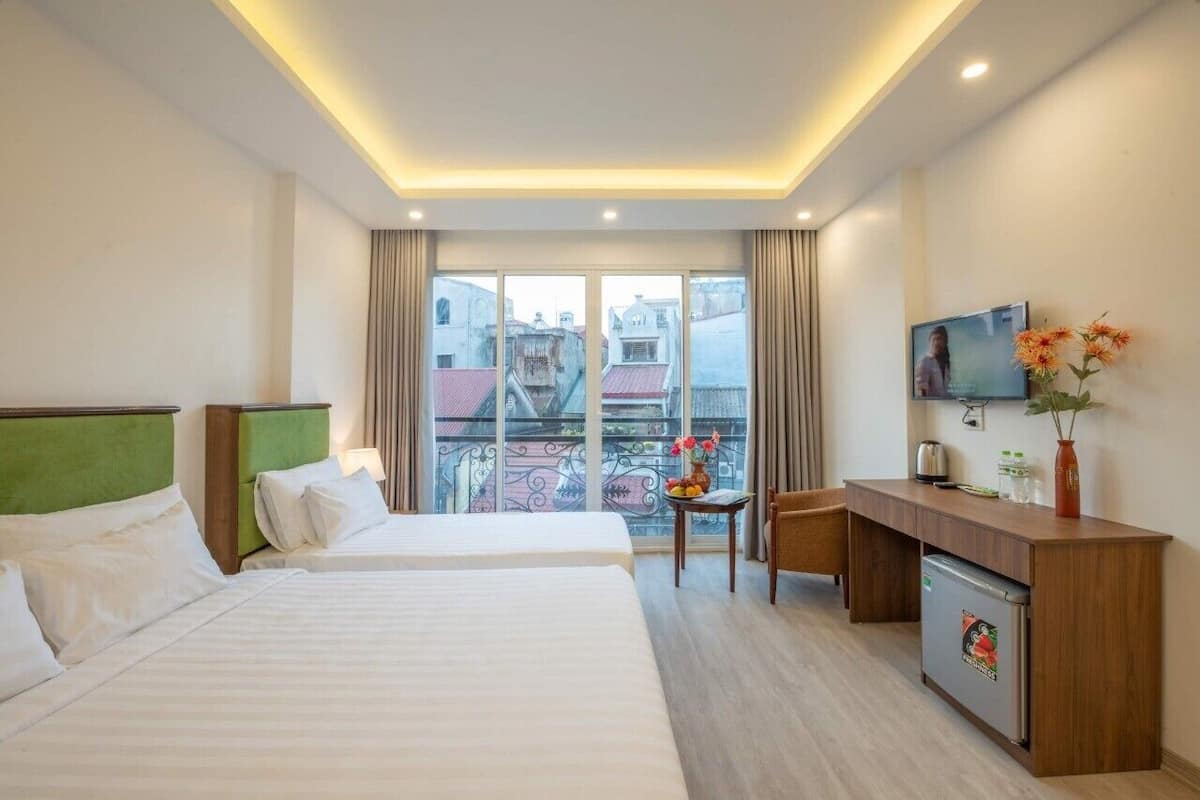 A hotel room with two beds, a desk, a TV, and a balcony with a view of buildings.