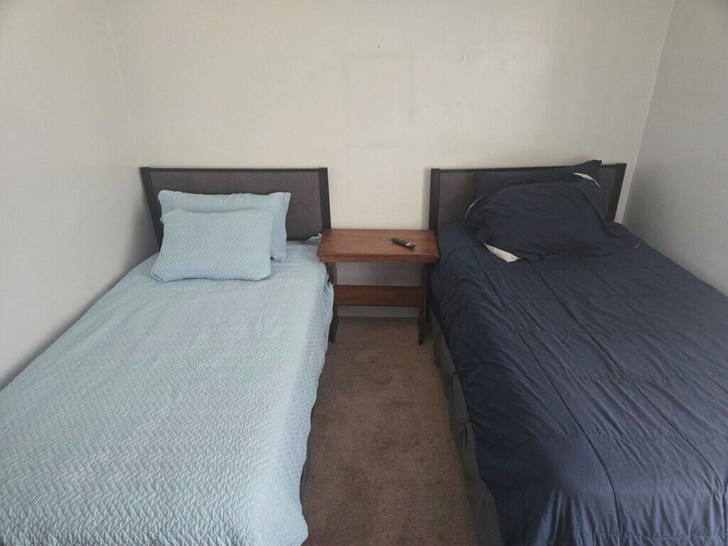 Een kleine hotelkamer met één ruimte, twee enkele bedden, een houten nachtkastje en een effen wand.