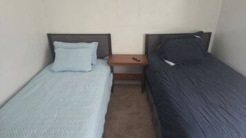Een kleine hotelkamer met één ruimte, twee enkele bedden, een houten nachtkastje en een effen wand.