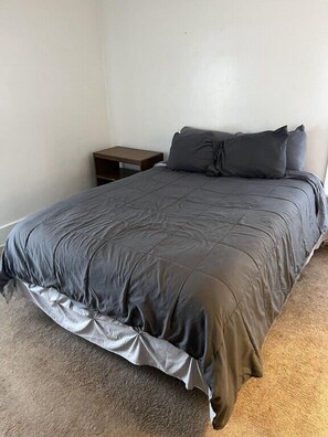 A single bed with a grey duvet and pillows, a wooden nightstand, and a carpeted floor.