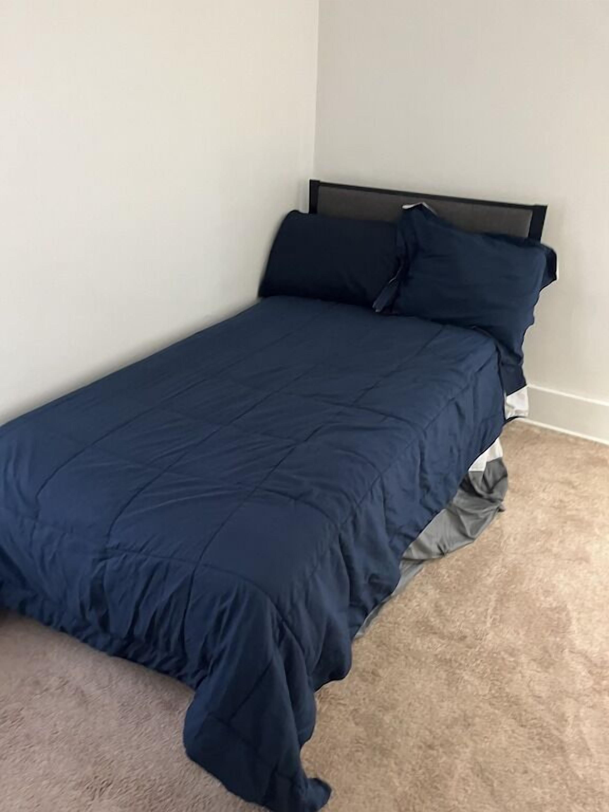 A single bed with a dark blue bedspread and matching pillows in a room with white walls and a beige carpet.