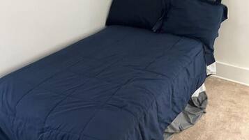 A single bed with a dark blue bedspread and matching pillows in a room with white walls and a beige carpet.