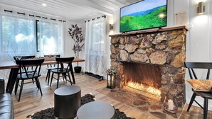 Un salon moderne avec une cheminée en pierre, un téléviseur à écran plat fixé au-dessus, et un coin repas comprenant des chaises et une table.