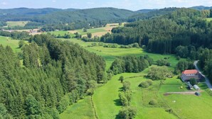 Paisagem rural com uma casa de campo, campos e colinas arborizadas.