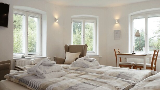 Quarto com cama grande, duas janelas, uma cadeira, uma mesa e um radiador.
