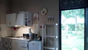 Een kleine keuken met witte kasten, een spoelbak, een kookplaat, een magnetron en een koelkast. Er is een raam met uitzicht op groen.