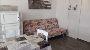 Een kamer met een houten vloer, een witte bank met een gestreept kussen, een witte tafel met een tafelkleed met koffieprint, een witte kast en een deur voorzien van haken.